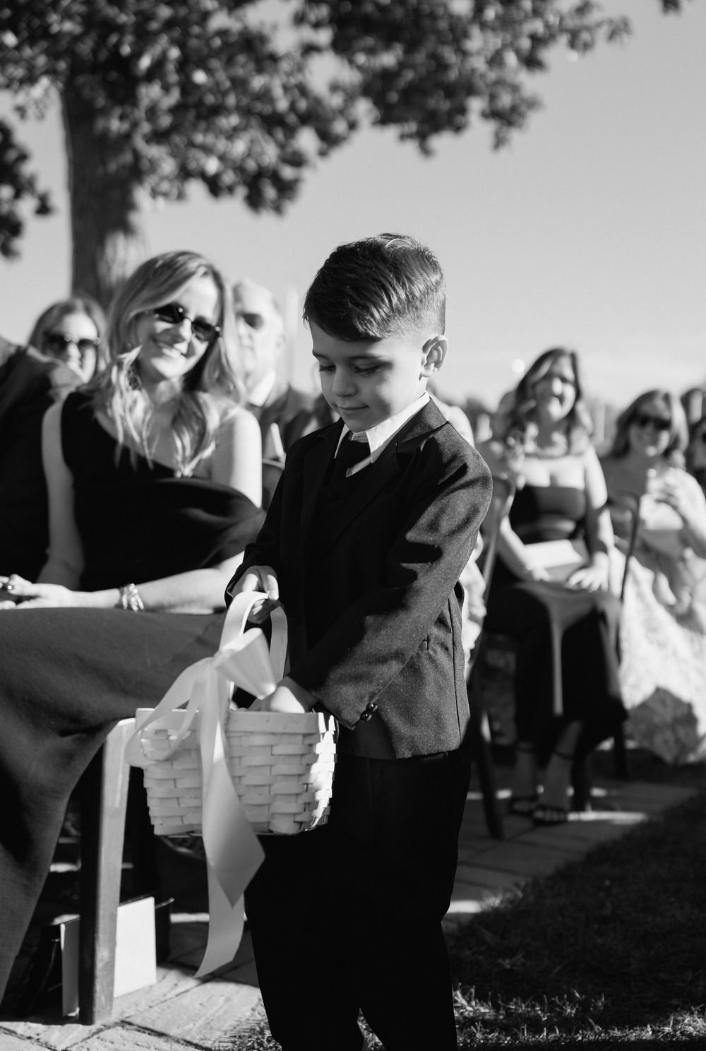 A young ring bearer in a suit carries a white basket with ribbon at an outdoor wedding ceremony.
