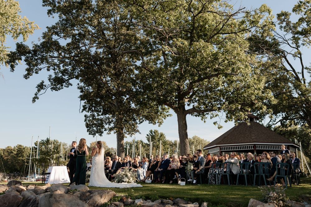 Outdoor wedding ceremony on a sunny day with guests seated near a gazebo under large trees on a lush green lawn.