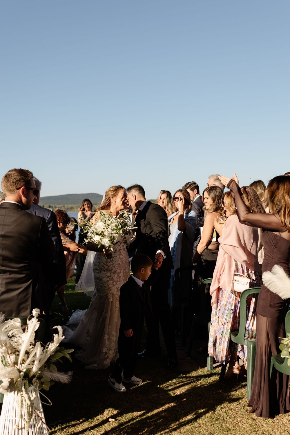 Bride and groom share a kiss surrounded by cheering wedding guests outdoors near a waterfront on a sunny day.