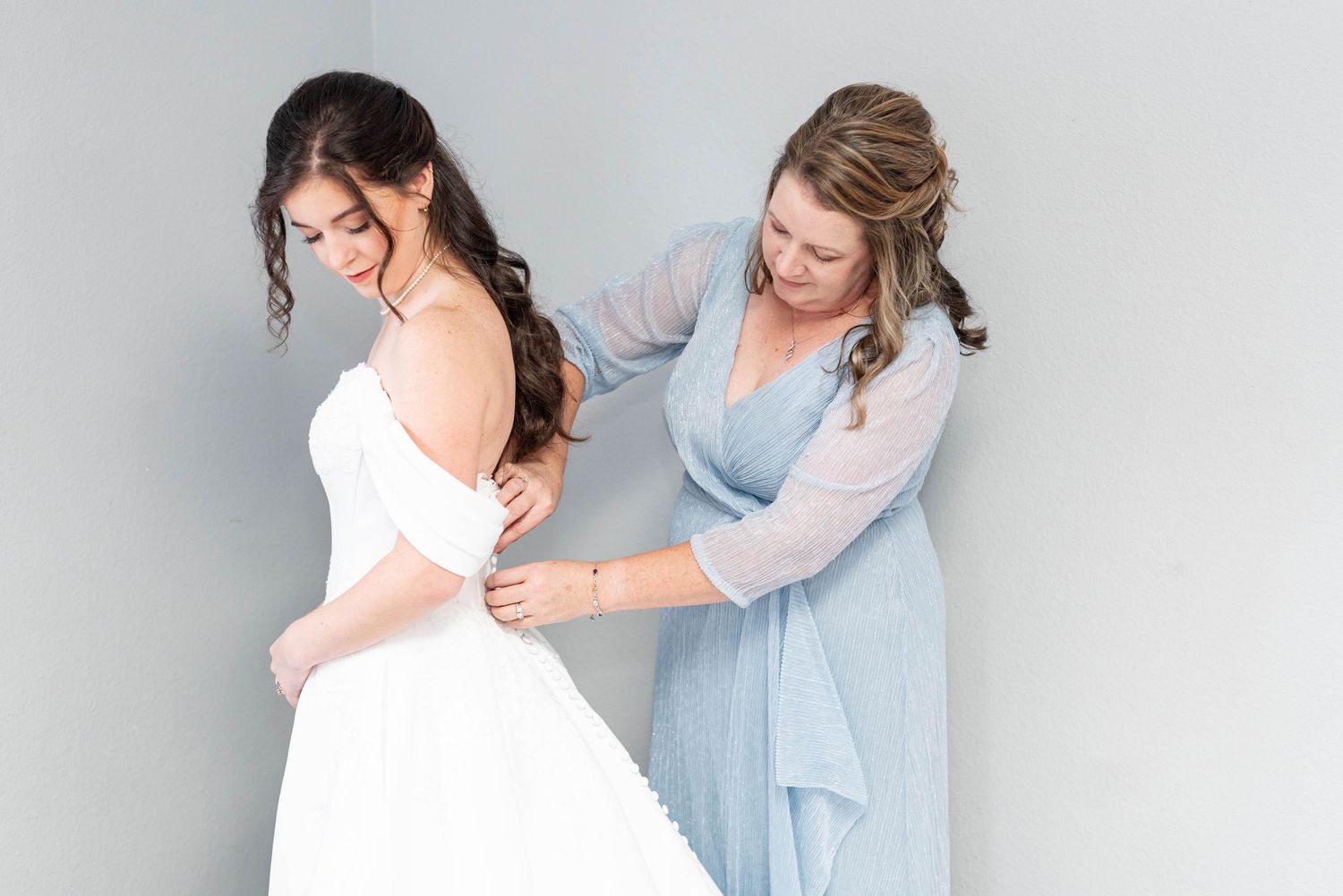 Bridesmaid in blue dress helping bride button up her white wedding gown before the ceremony.