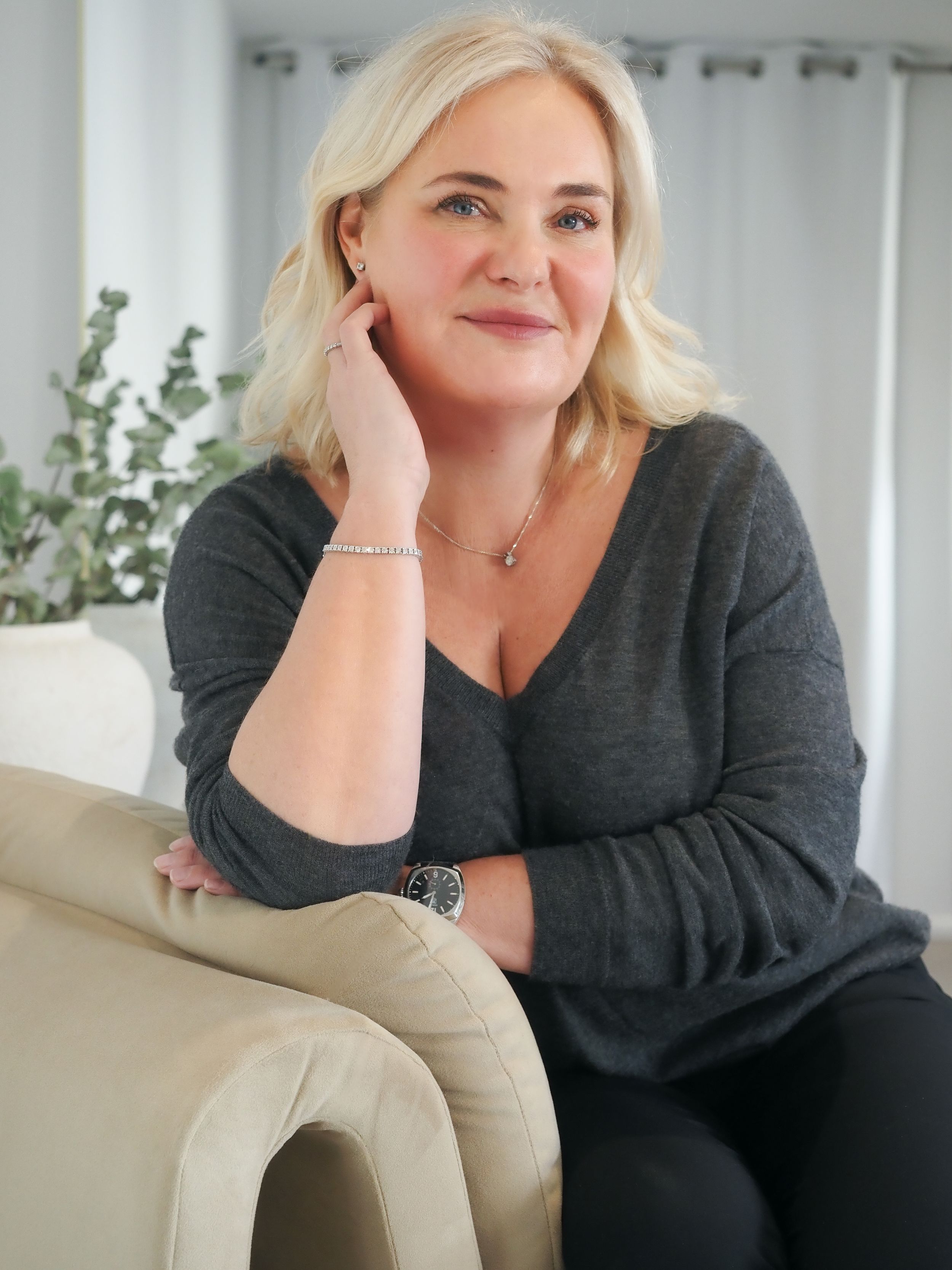 Blonde woman in gray sweater sitting relaxed on beige chair, wearing watch and bracelet, smiling softly.