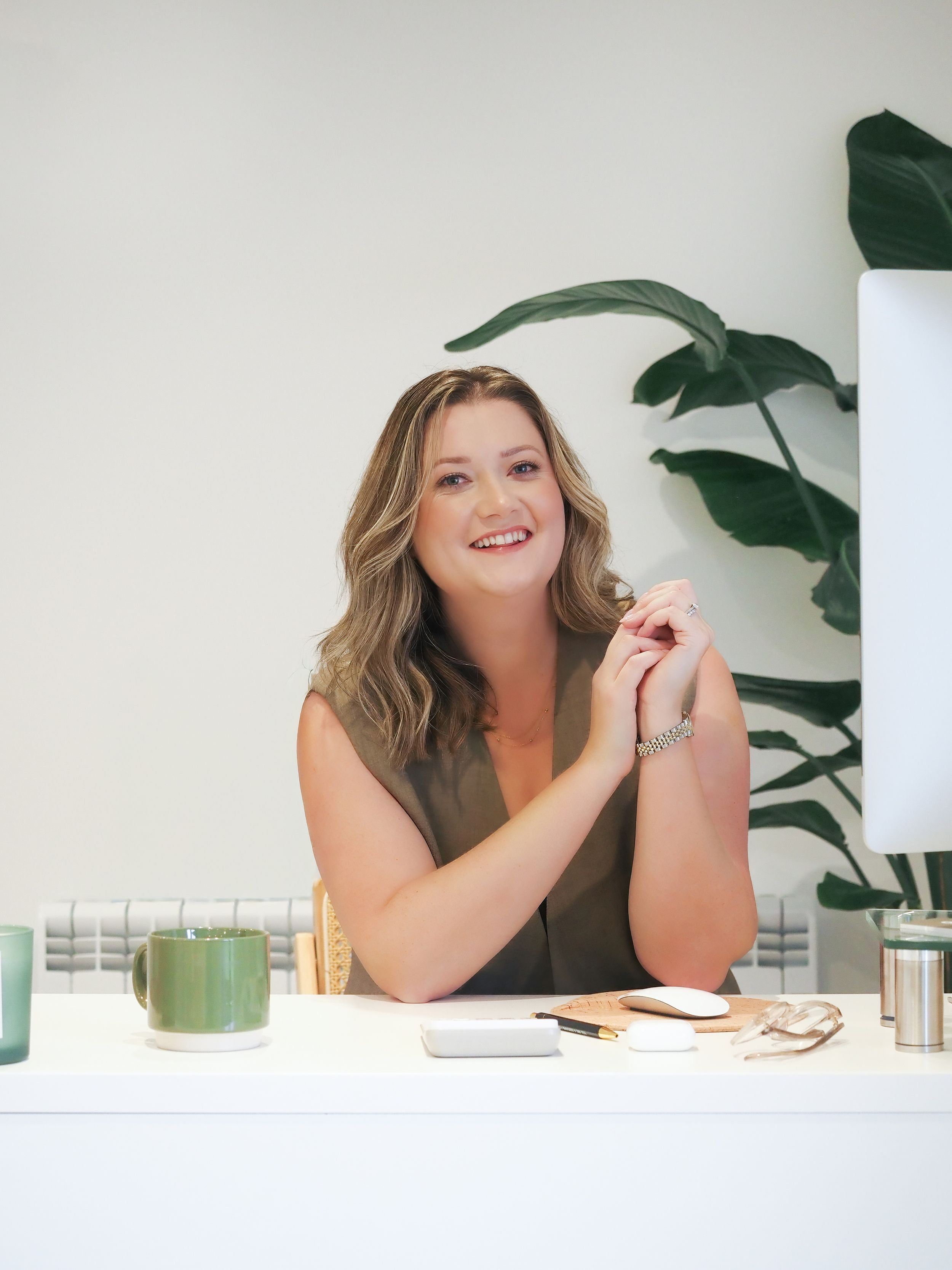 Smiling woman sitting at a white desk with green mug and plant, wearing olive top in bright modern office.