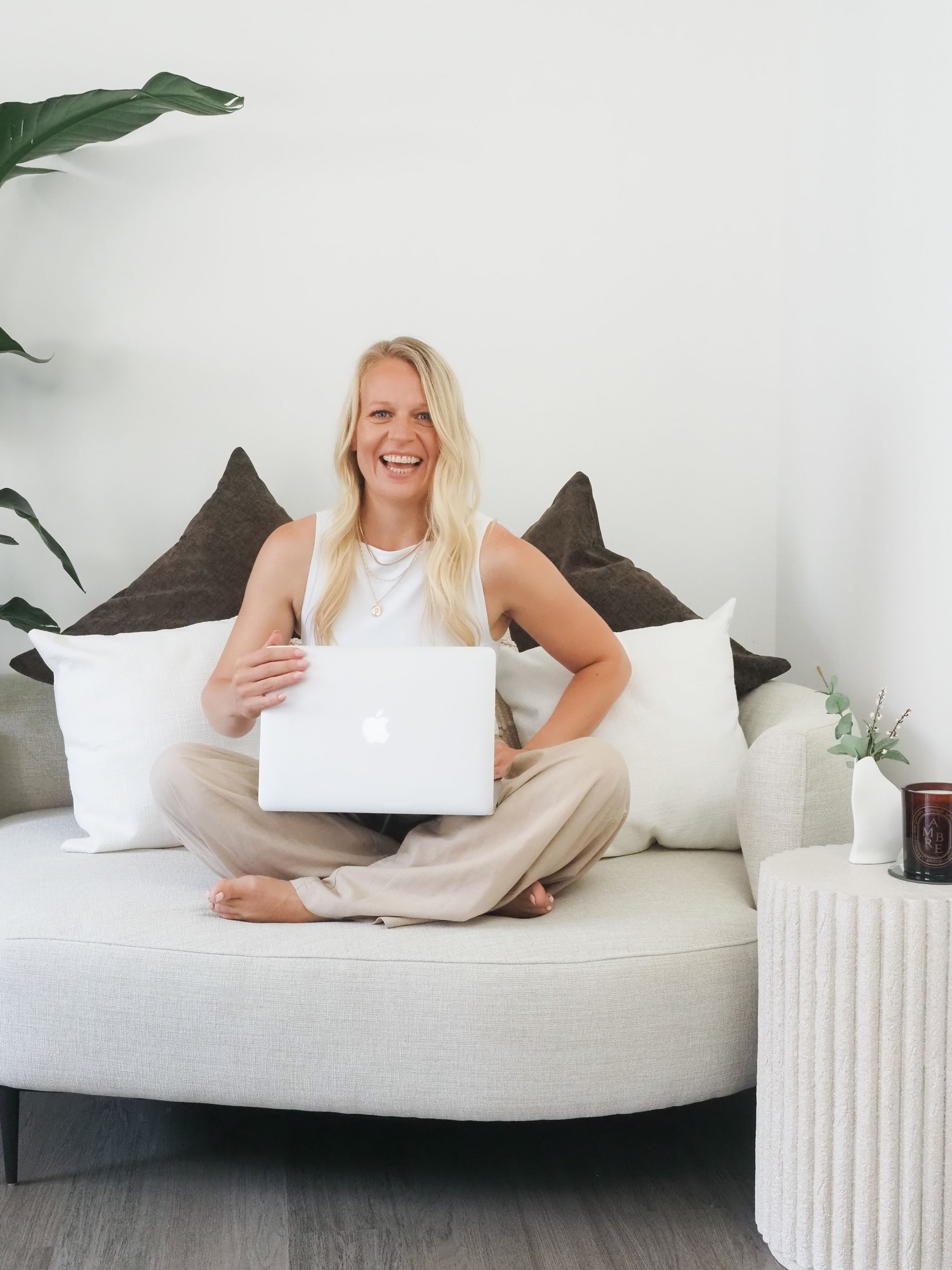 Smiling blonde woman sitting cross-legged on white sofa holding MacBook laptop in bright modern living room.
