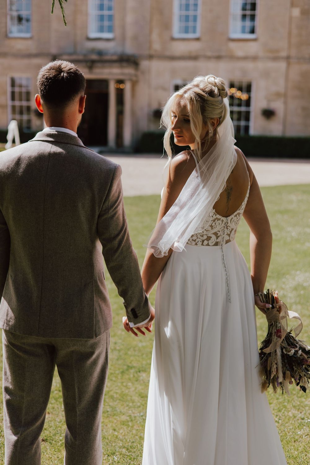 Bride in lace wedding dress with veil holds groom's hand in gray suit walking toward elegant manor house.