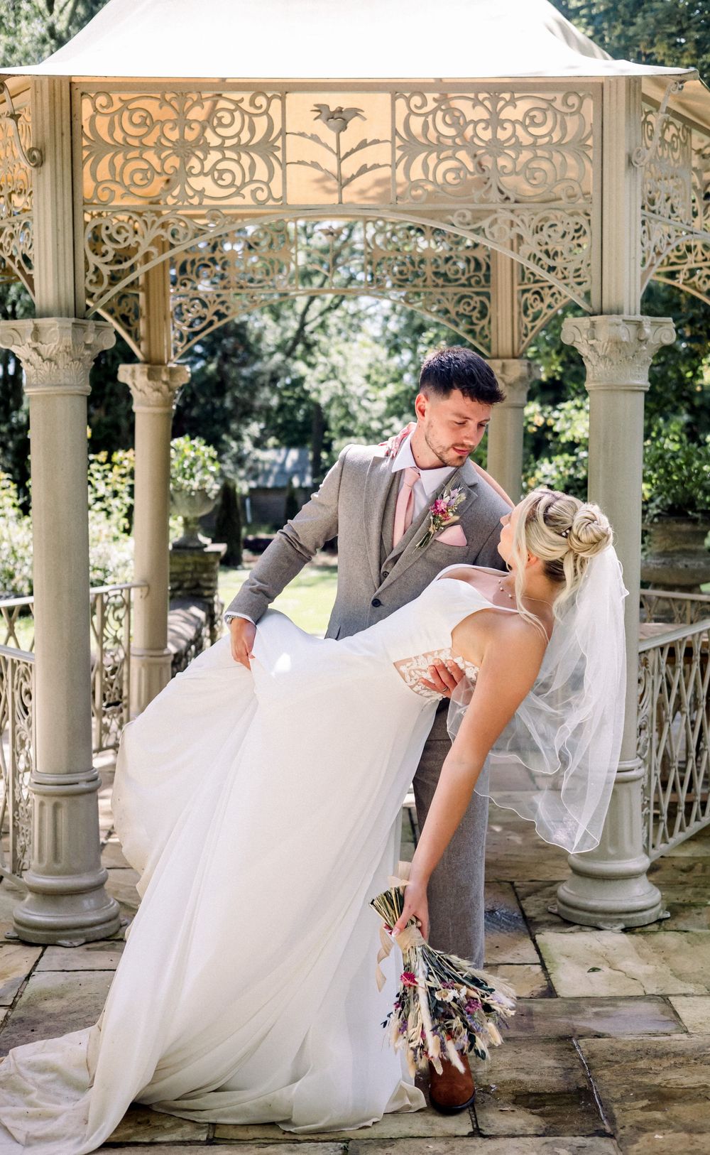 A bride and groom share a romantic dip kiss under an ornate white gazebo in a lush garden setting.
