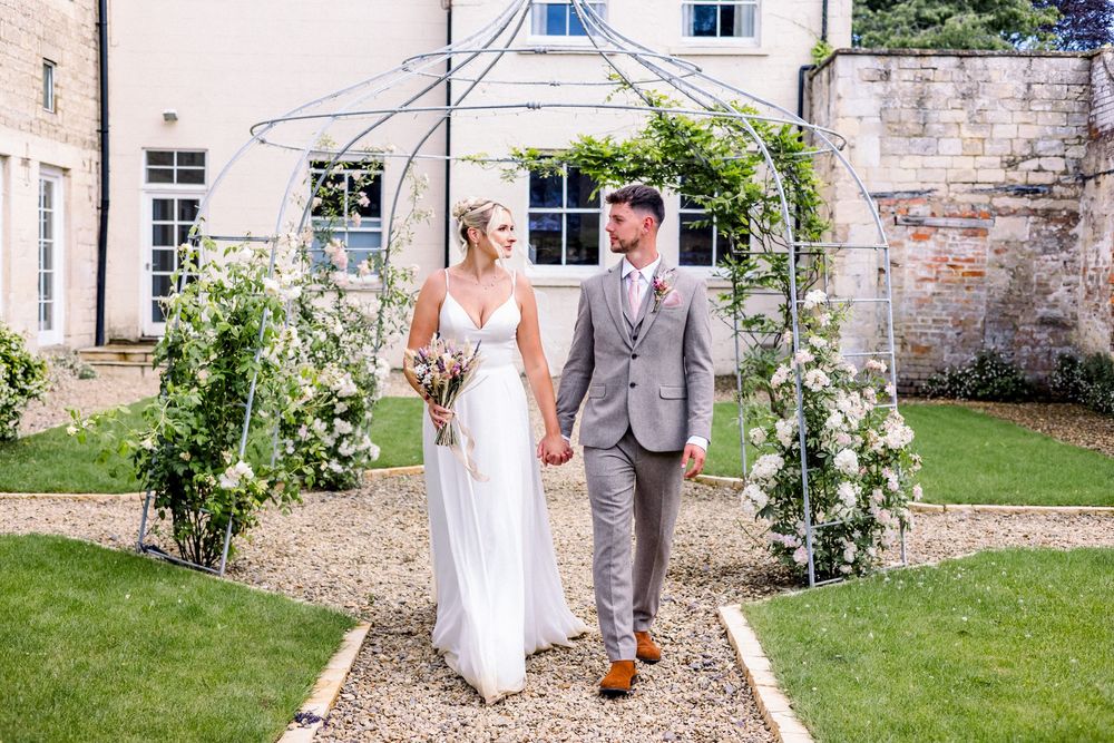 Bride and groom walk hand in hand under a floral arch in a sunny garden at an elegant countryside wedding venue.