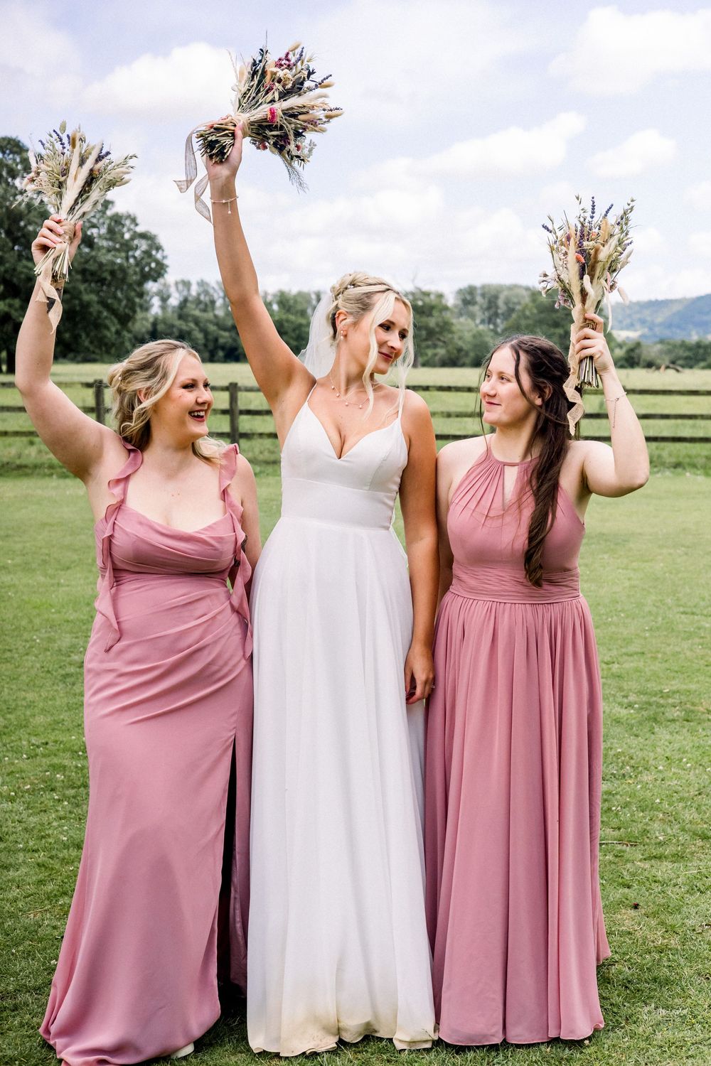Bride in white gown with two bridesmaids in pink dresses raising bouquets outdoors at a farm wedding.