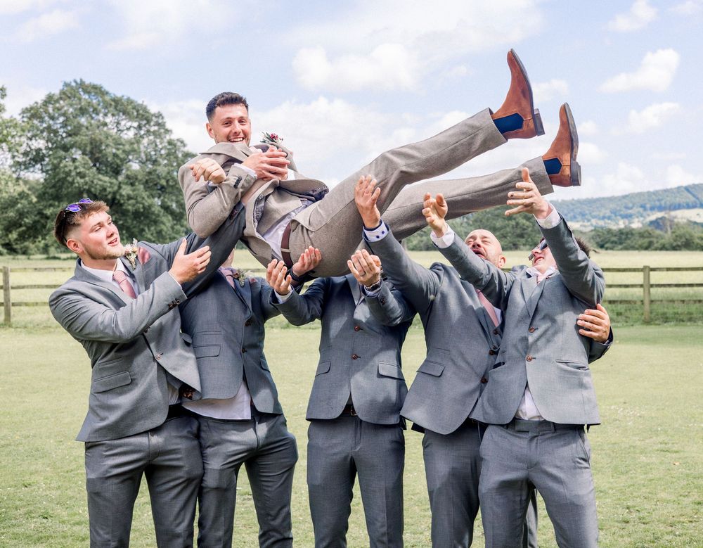 Groomsmen in gray suits lifting the groom in the air outdoors at a wedding celebration.
