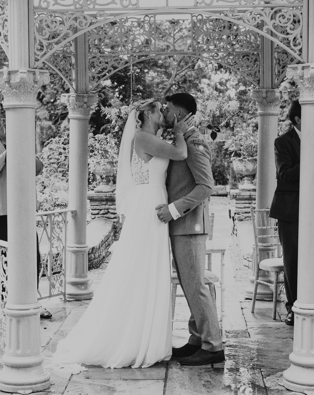 Black and white photo of a bride and groom sharing their first kiss under an ornate white gazebo.
