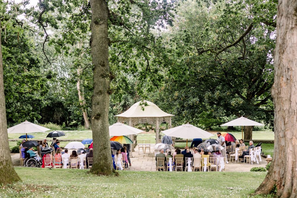 Outdoor wedding ceremony in lush green park with guests holding colorful umbrellas seated around a white gazebo altar.