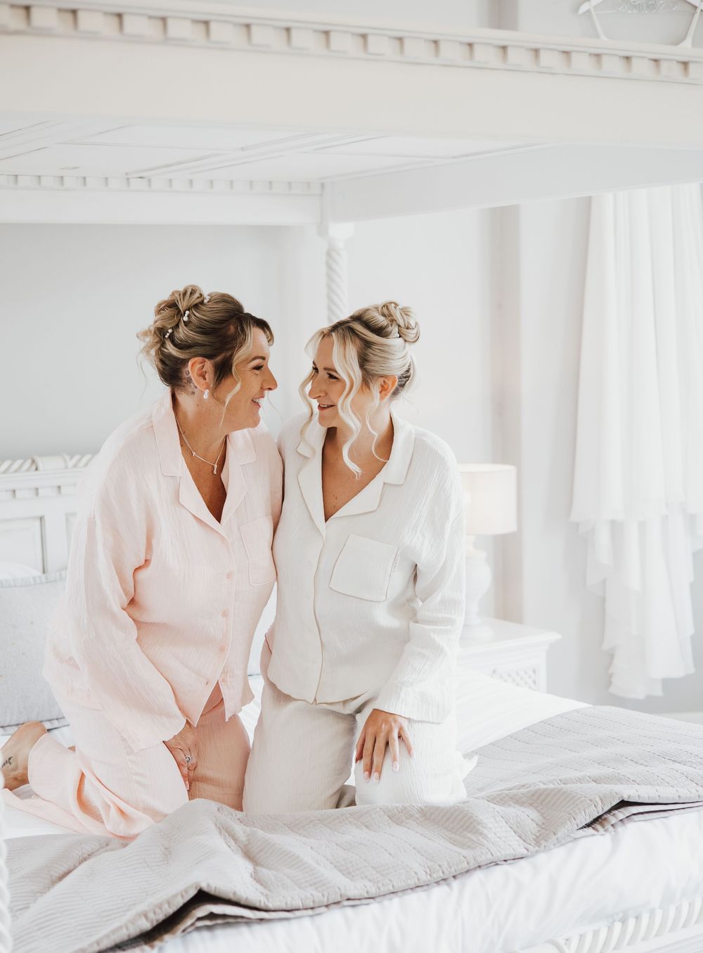 Two women in matching pajamas sit on a white bed, smiling at each other with elegant updo hairstyles.