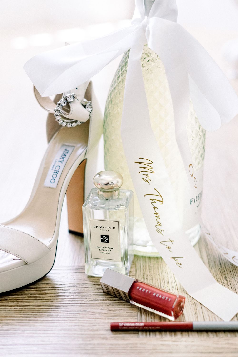 Bridal flat lay featuring Jimmy Choo heels, Jo Malone perfume, Fenty Beauty lip liner, and a personalized wedding bag.
