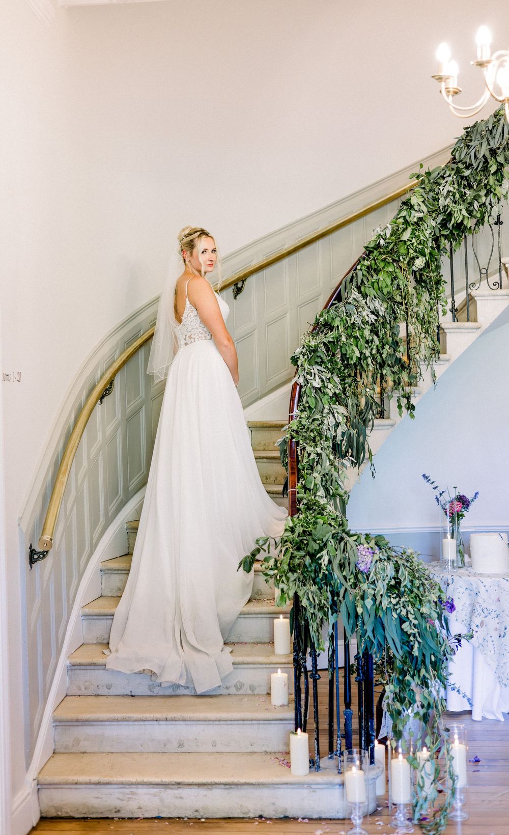 Bride in white gown poses on elegant staircase adorned with lush greenery garland and candles at wedding venue.