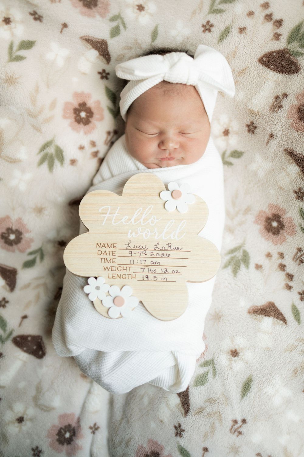 Newborn baby girl swaddled in white with bow headband, holding a wooden Hello World birth announcement sign.