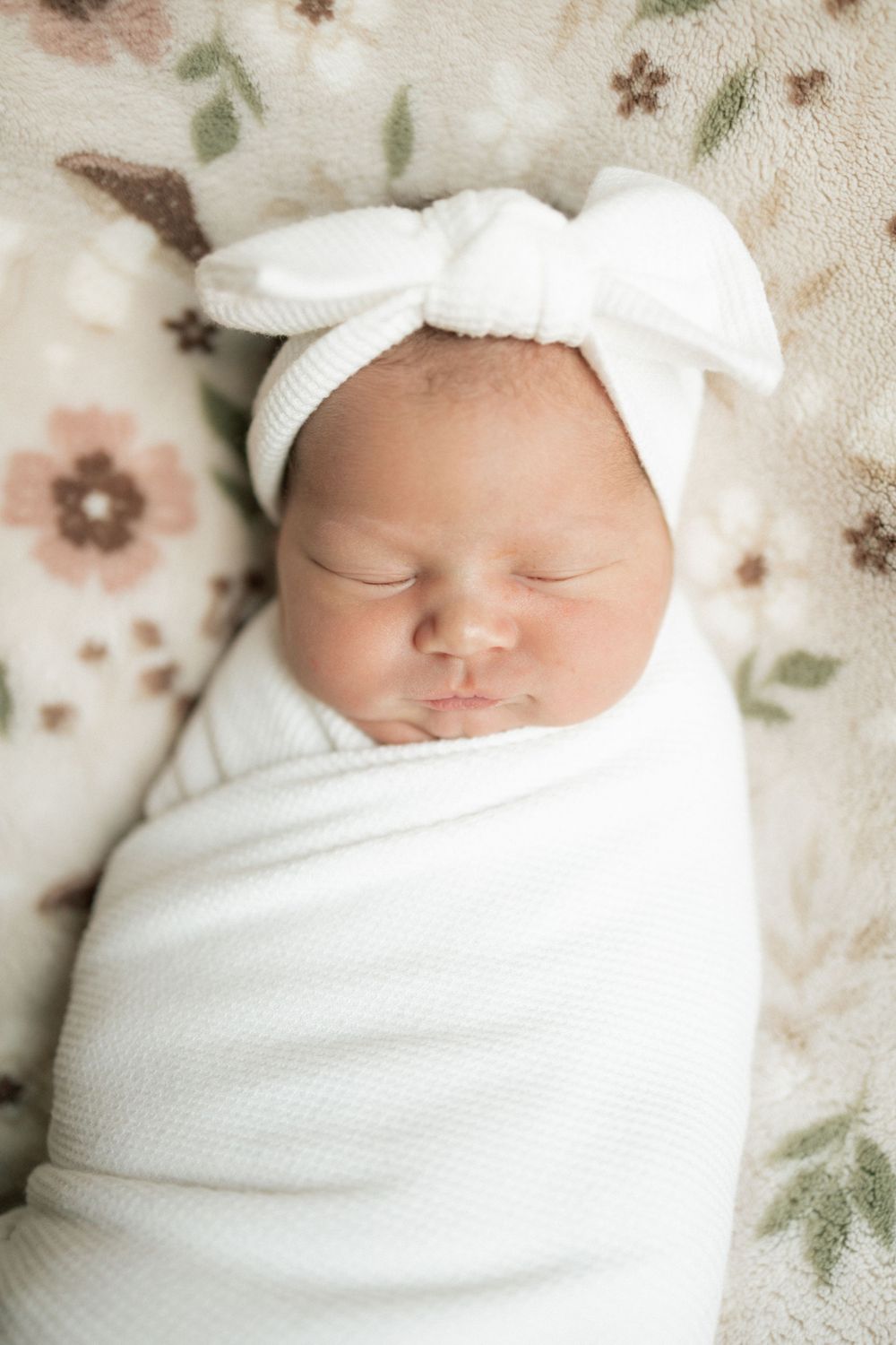Sleeping newborn baby swaddled in white wrap with bow headband on floral blanket.