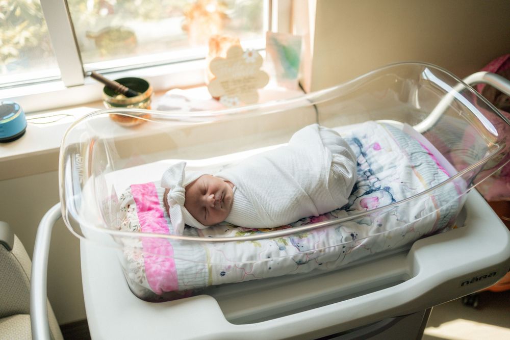 Newborn baby swaddled in white blanket resting peacefully in a hospital bassinet by a sunny window.