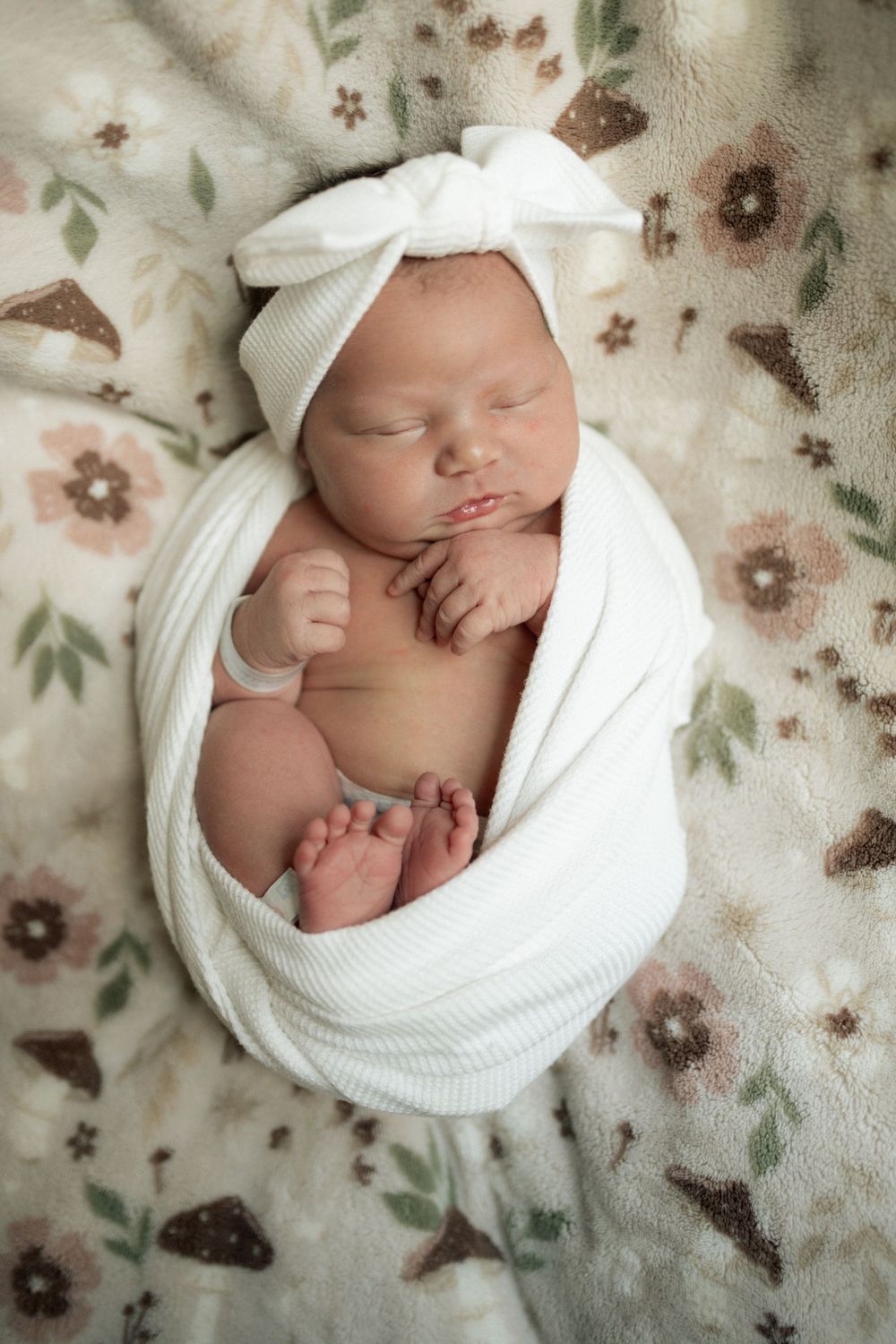 Newborn baby girl swaddled in white wrap with bow headband, sleeping peacefully on floral blanket.