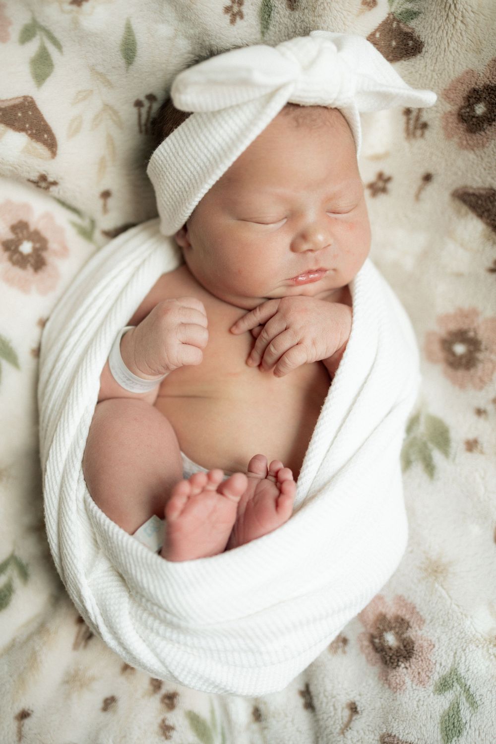 Sleeping newborn baby girl swaddled in white wrap with bow headband on floral blanket.
