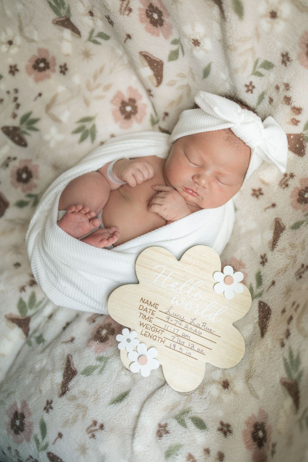 Newborn baby girl in white swaddle and bow headband beside wooden birth announcement sign on floral blanket.