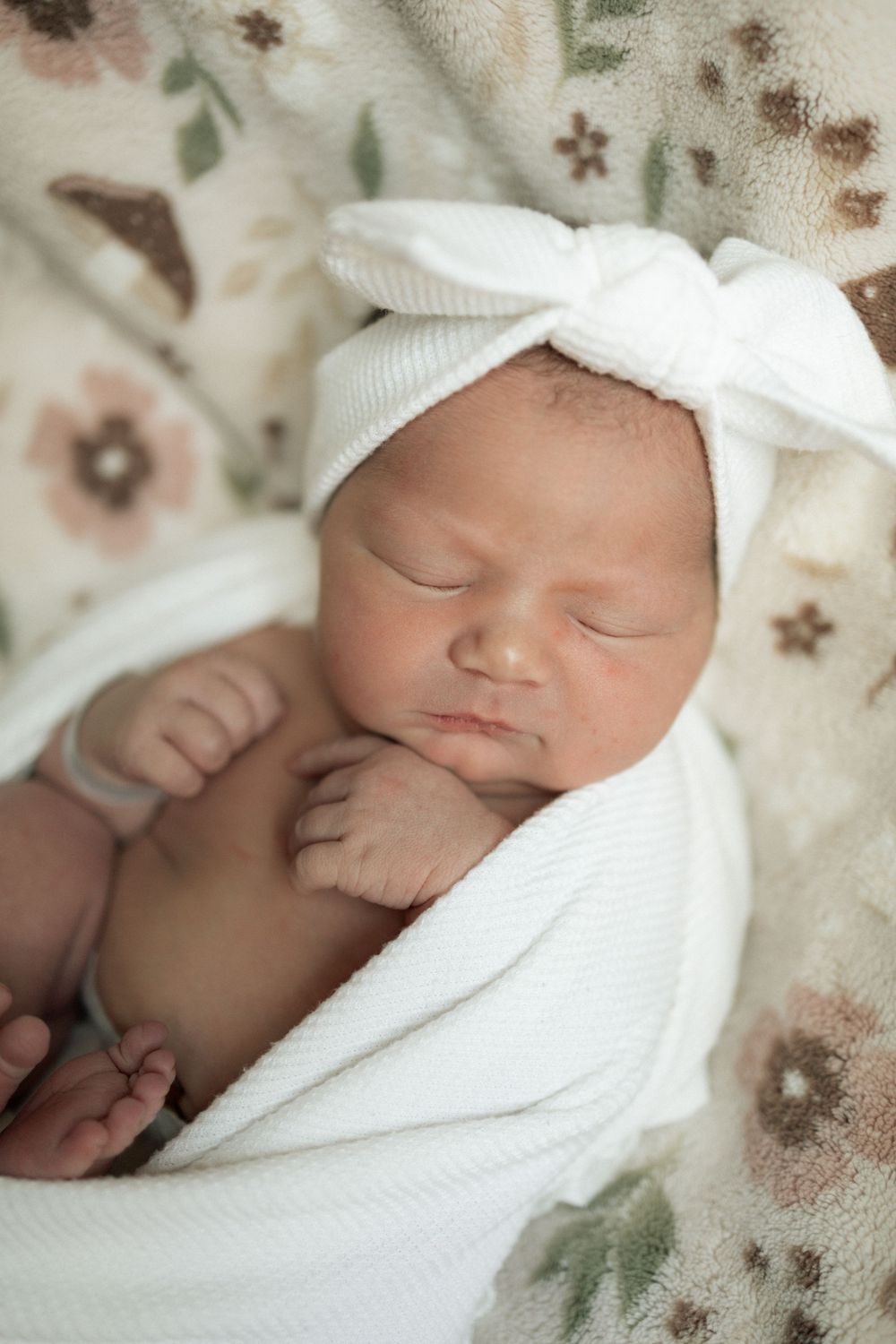 Sleeping newborn baby wrapped in white swaddle with bow headband on floral blanket.