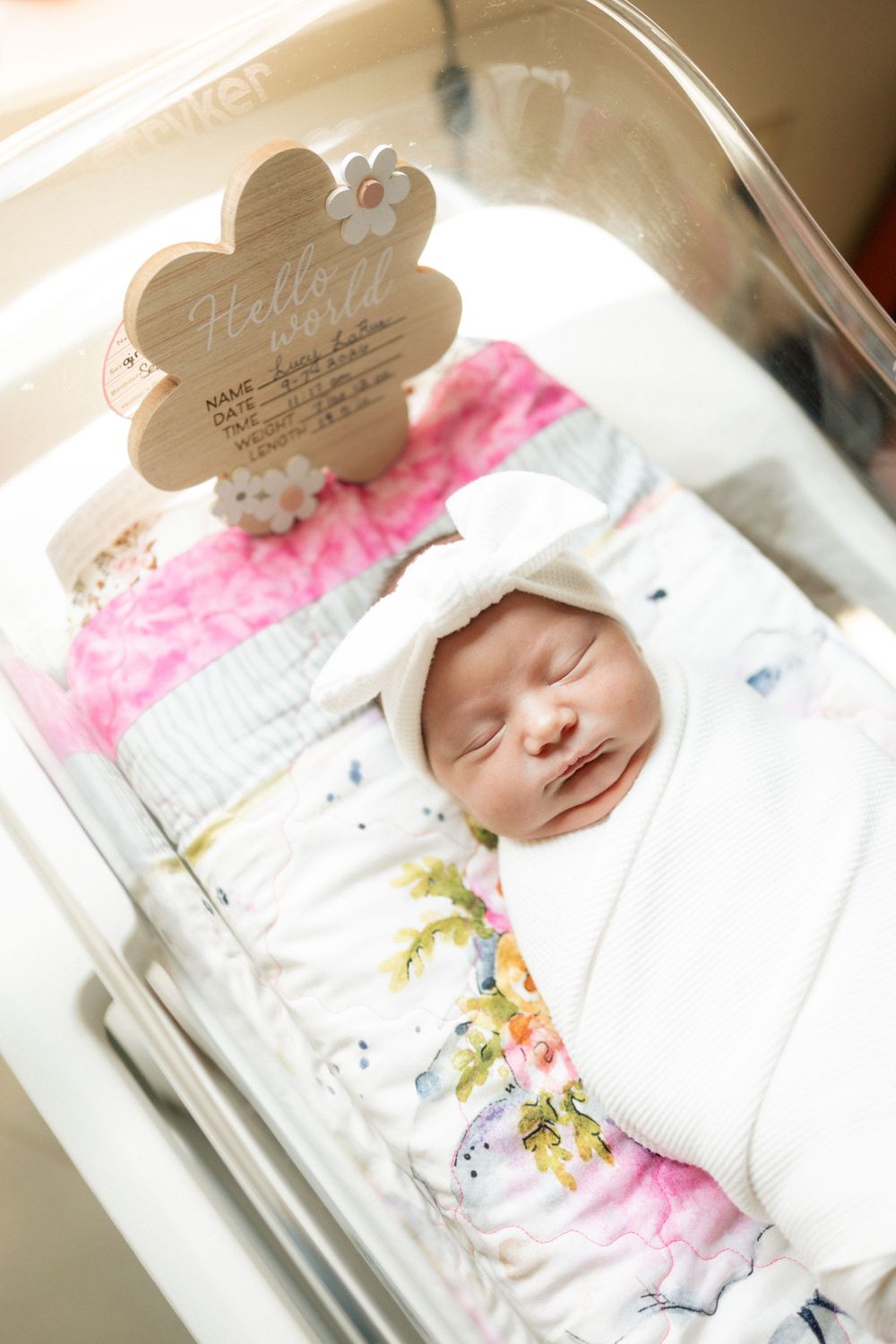 Newborn baby girl swaddled in floral blanket sleeping in hospital bassinet with Hello World wooden sign.