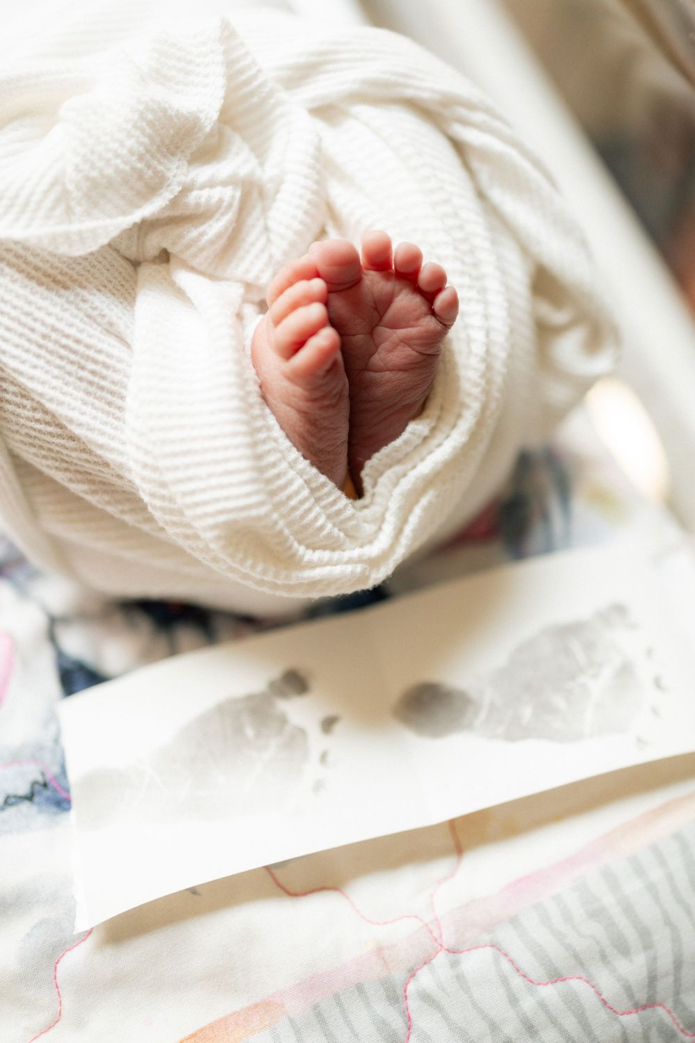 Close-up of newborn baby feet peeking out of a white swaddle blanket next to footprint card in hospital.