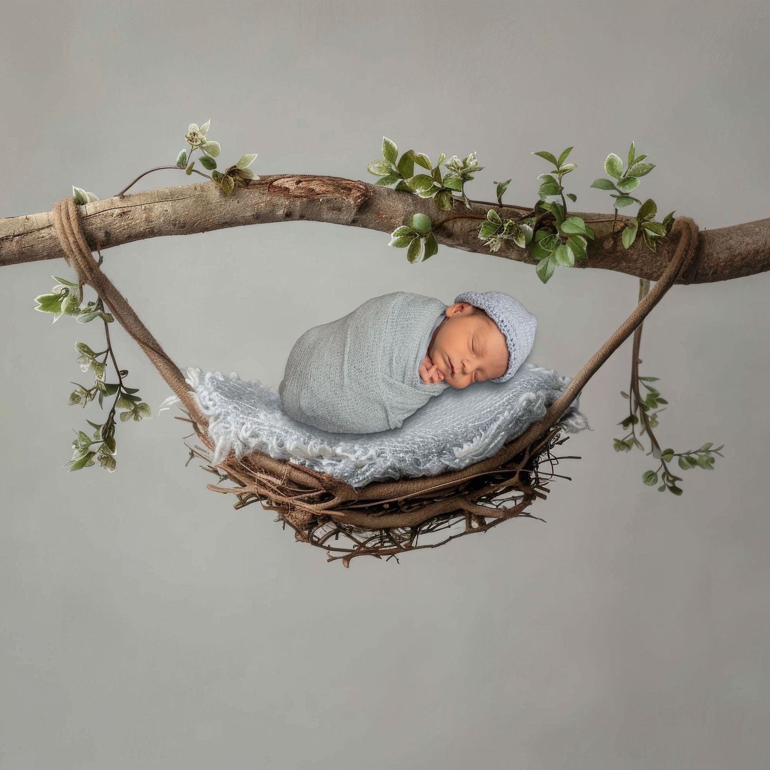 Newborn baby swaddled in blue wrap sleeping peacefully in a rustic twig nest hammock with white flowers.