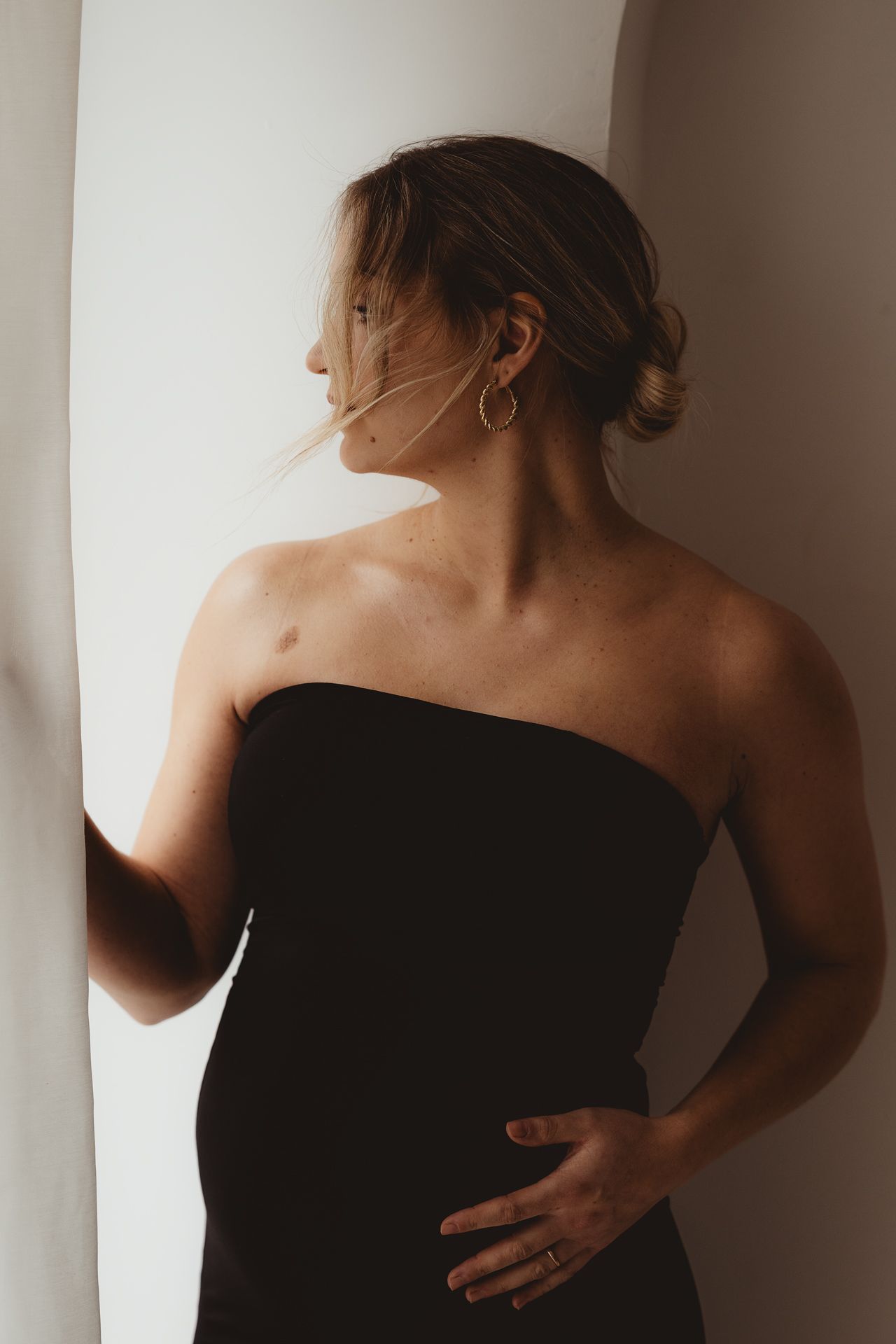 Pregnant woman in black strapless dress poses barefoot in photography studio with professional lighting equipment.