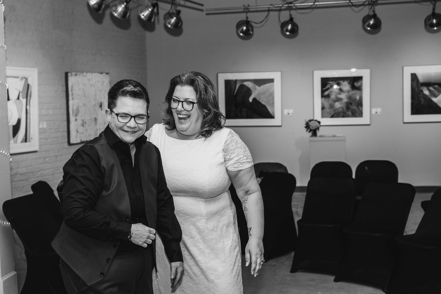 Black and white photo of two people smiling together in an art gallery with framed photos on the wall.