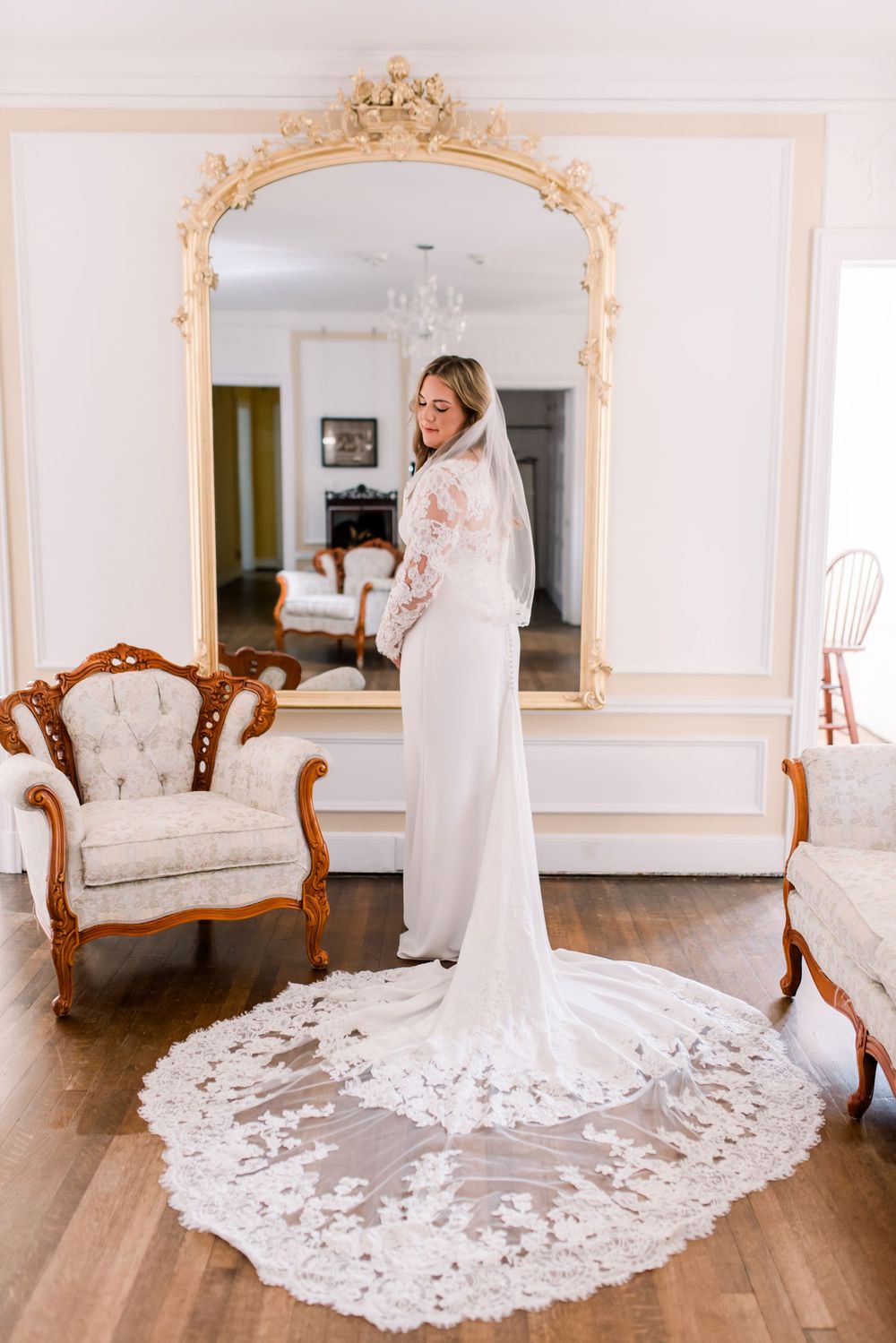 Bride in elegant white lace wedding gown with cathedral veil stands before ornate gold mirror in vintage-styled room.