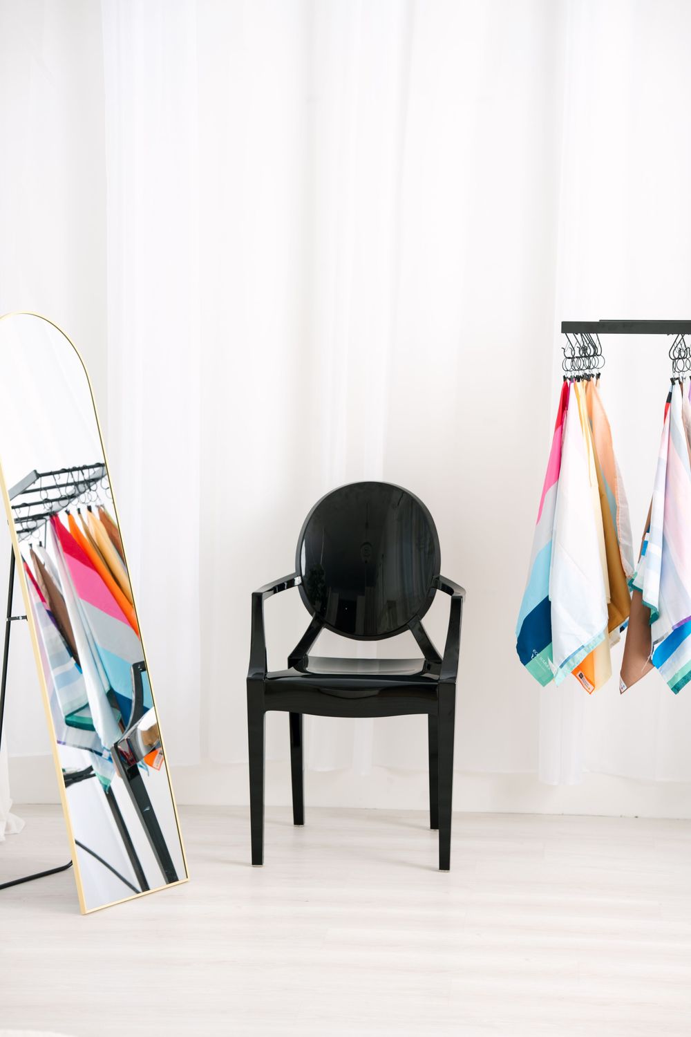 Black ghost chair between full-length mirror and colorful scarves on clothing rack.