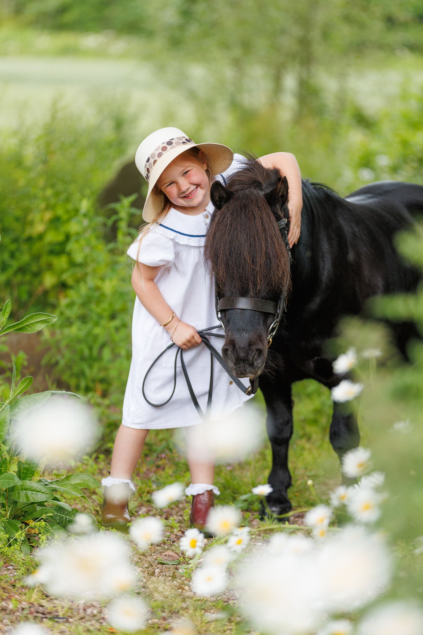 Young girl in white dress and hat smiling while hugging a black pony in a flower-filled meadow.