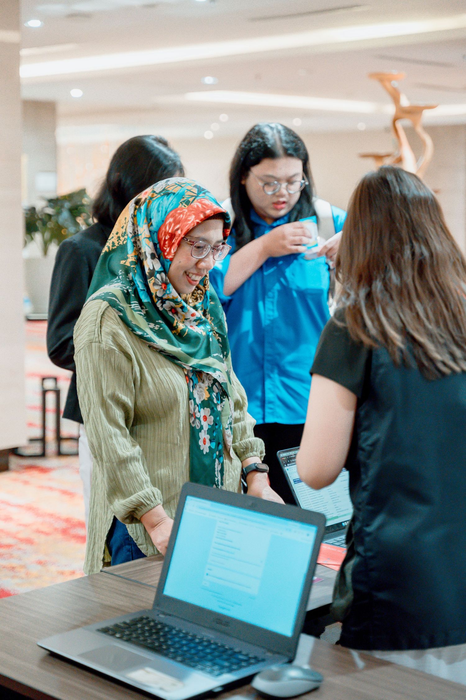Woman in colorful hijab smiling at hotel registration desk with laptop, assisted by staff at a professional event.