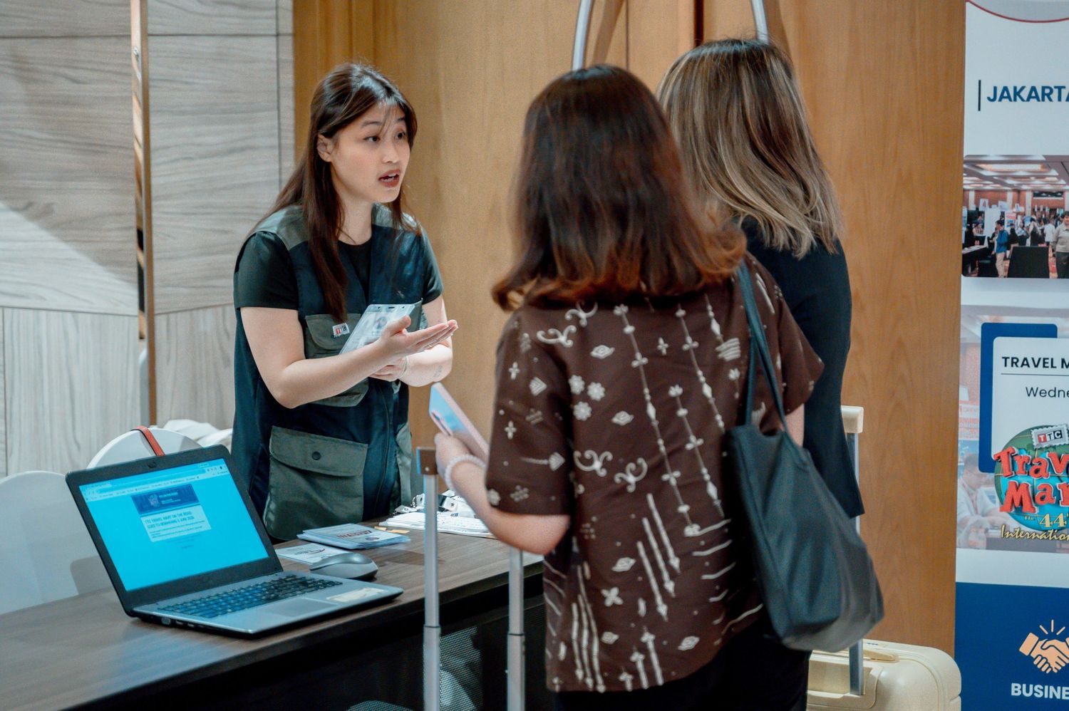 A woman at a registration desk assists two attendees at a business travel expo event in Jakarta.