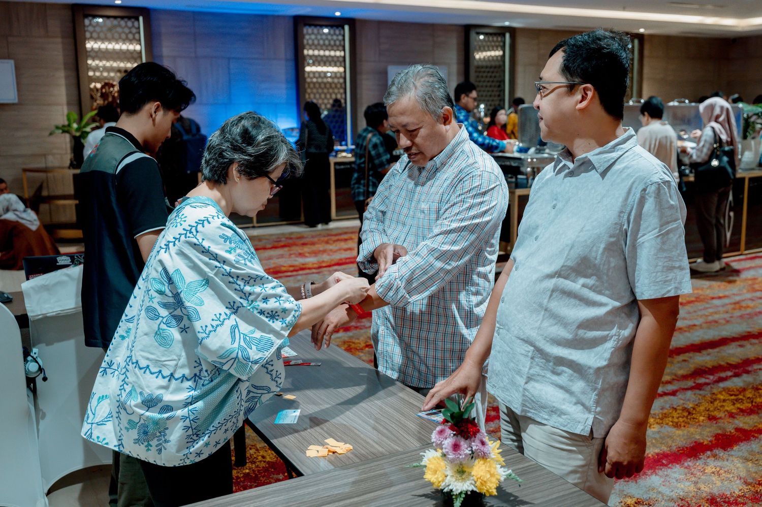 Woman in blue batik hands items to guests at a formal indoor event in an elegantly decorated conference hall.