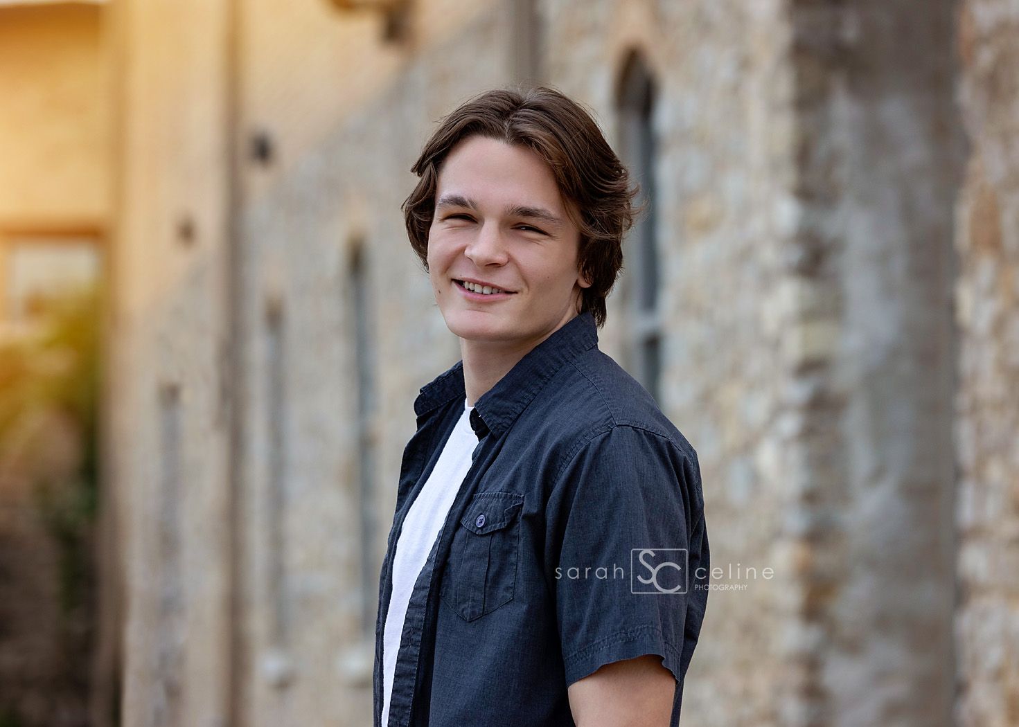 Smiling teenage boy with medium-length hair wearing a dark button-up shirt, posing outdoors near a stone building.