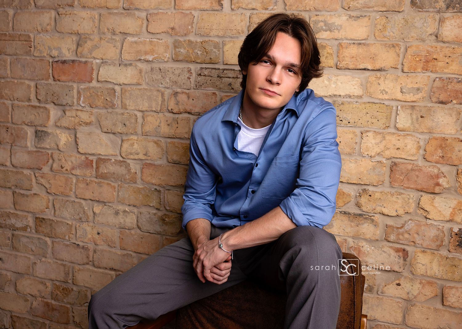 Young man in blue shirt sitting against a rustic brick wall in a professional portrait photo.