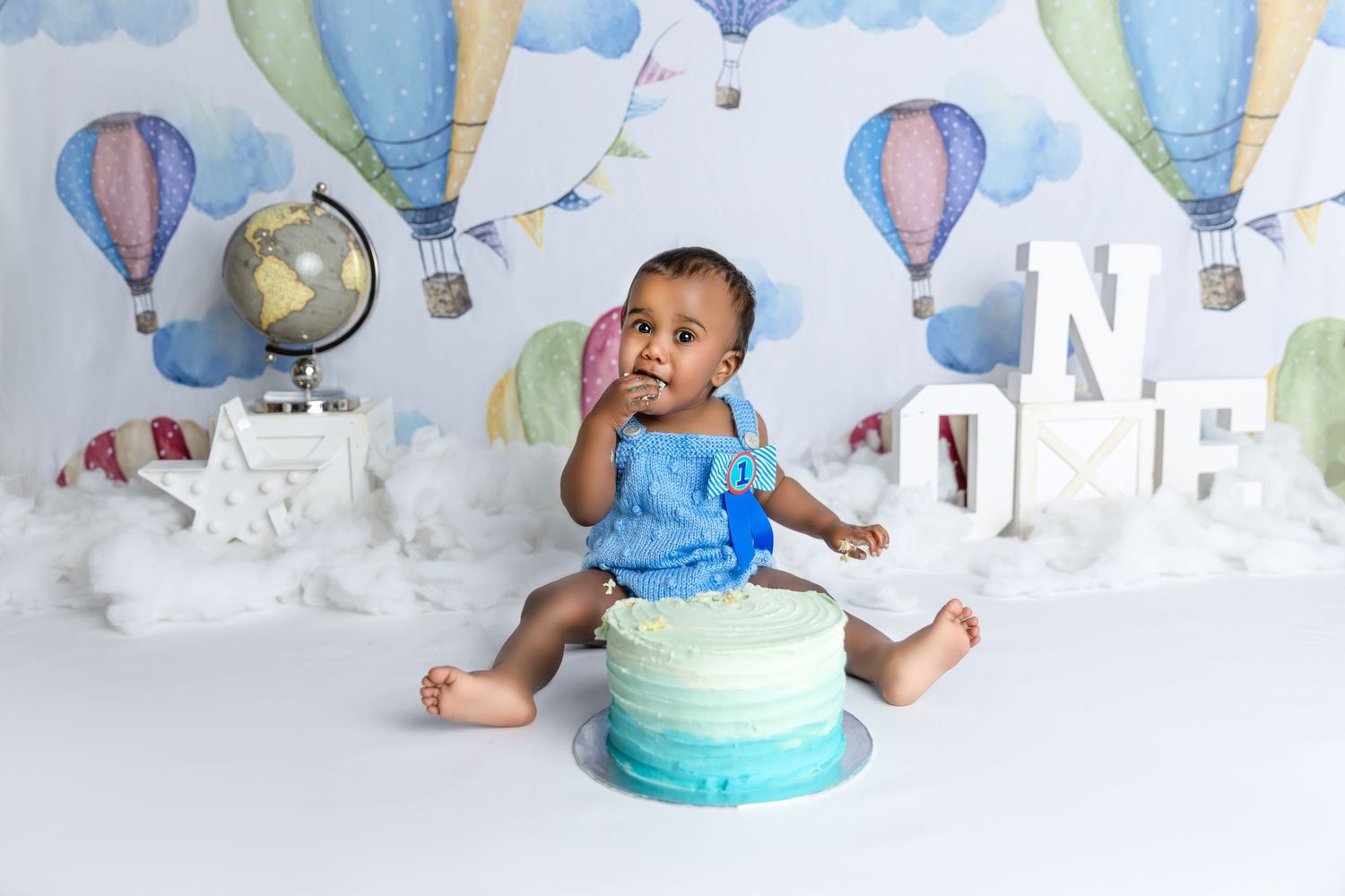 Baby's first birthday cake smash photo with hot air balloon backdrop, blue ombre cake, and ONE letters.