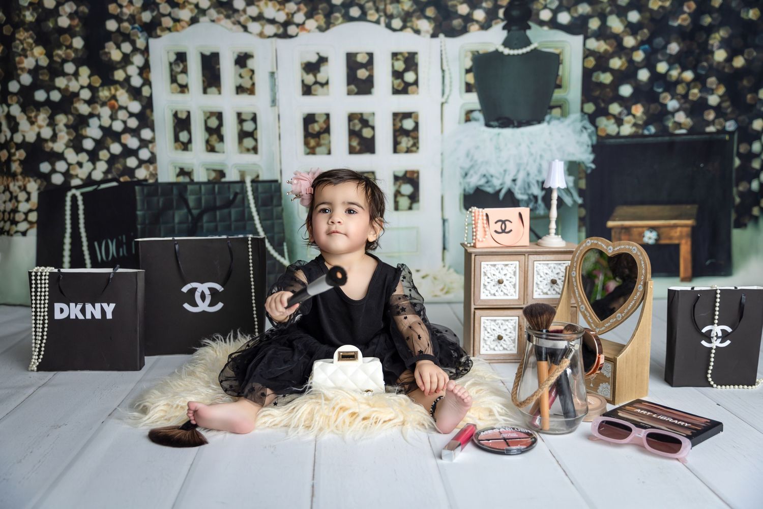 Toddler girl in black lace dress poses with Chanel and DKNY bags, makeup, and mirror in a fashion-themed photo shoot.