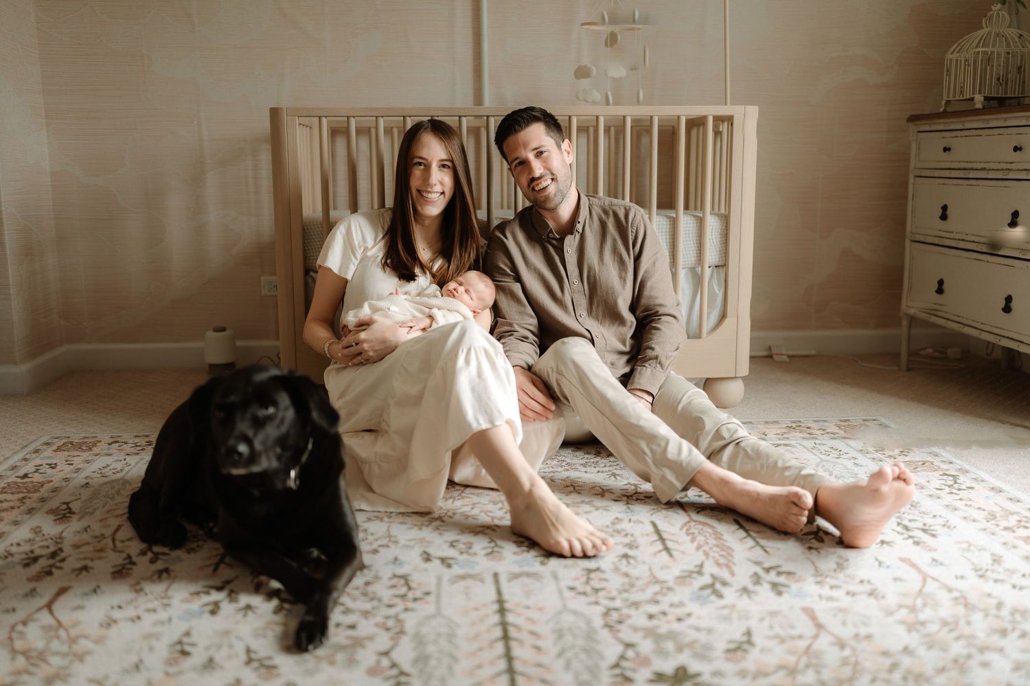 Couple sitting on nursery floor with newborn baby and black Labrador dog in front of white crib.