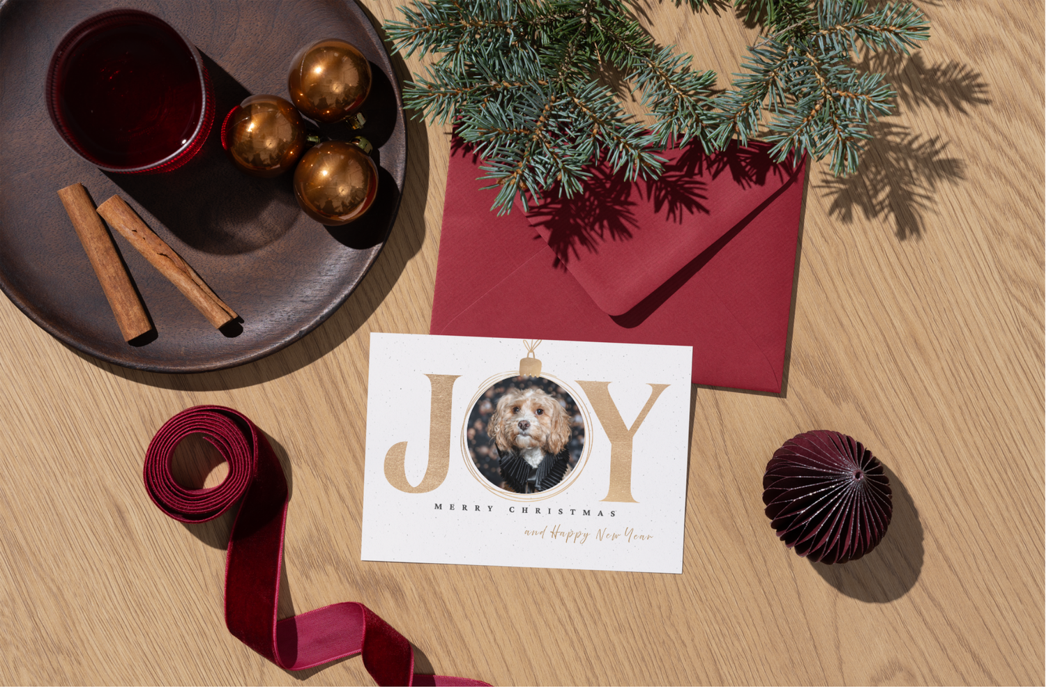 Gold and white JOY Christmas card with dog photo ornament, red envelope, pine branches, and holiday decorations on wood surface.