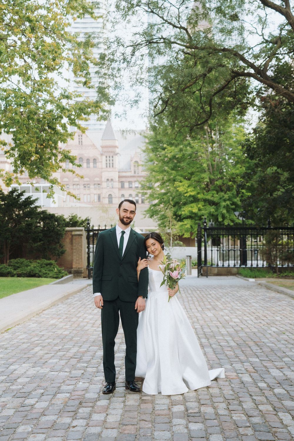 Bride and groom posing on cobblestone path with lush trees and historic brick building backdrop.