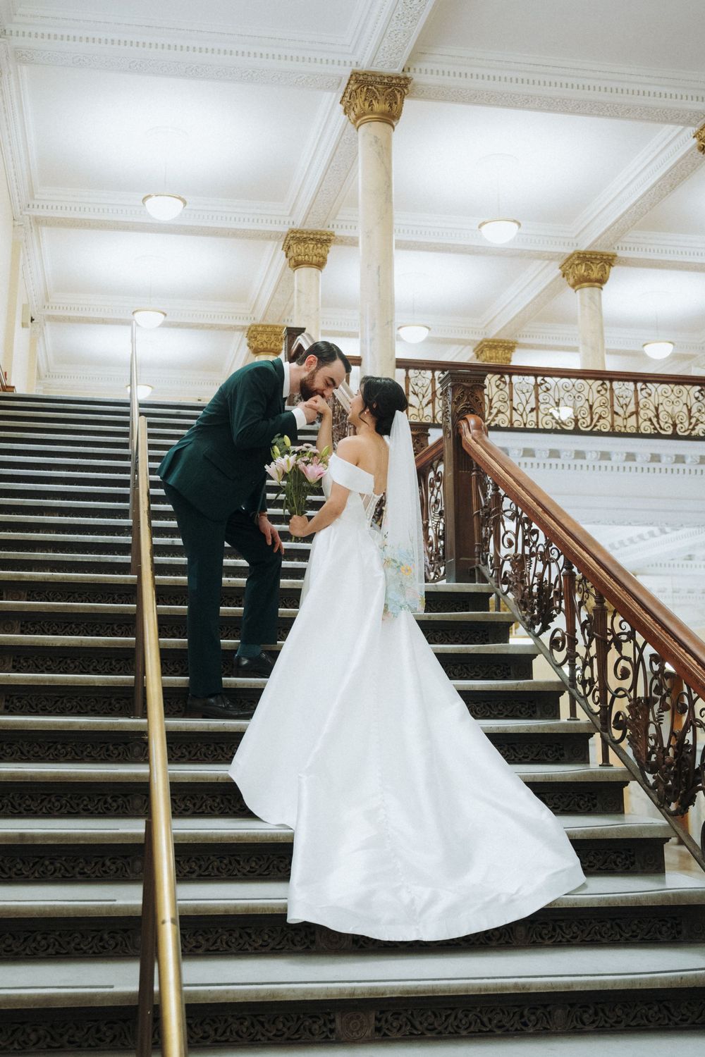 Bride and groom share a romantic moment on grand ornate staircase, bride in off-shoulder white gown, groom in dark suit.