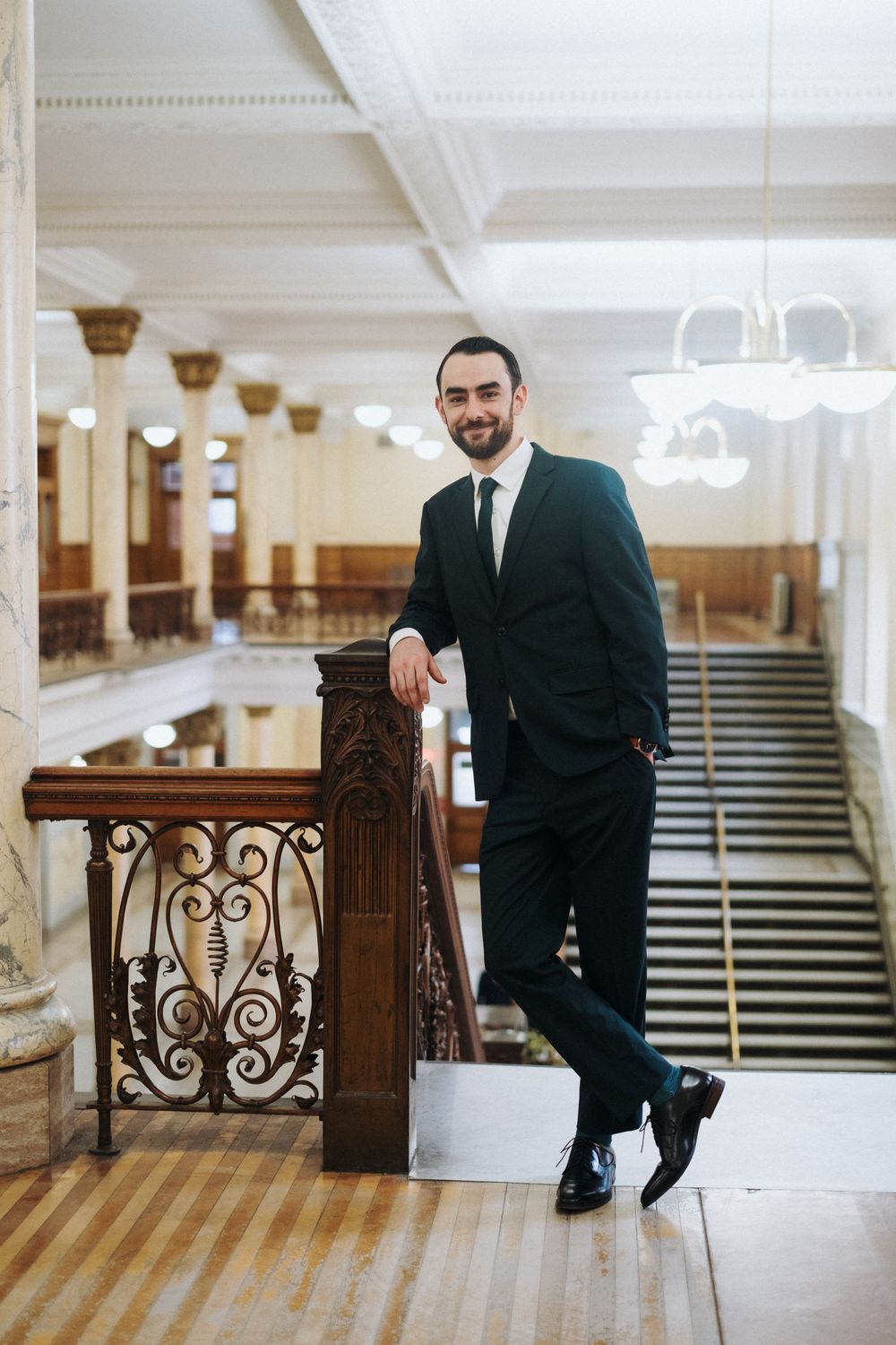 Smiling bearded man in black suit leaning on ornate railing in elegant grand hall with columns.