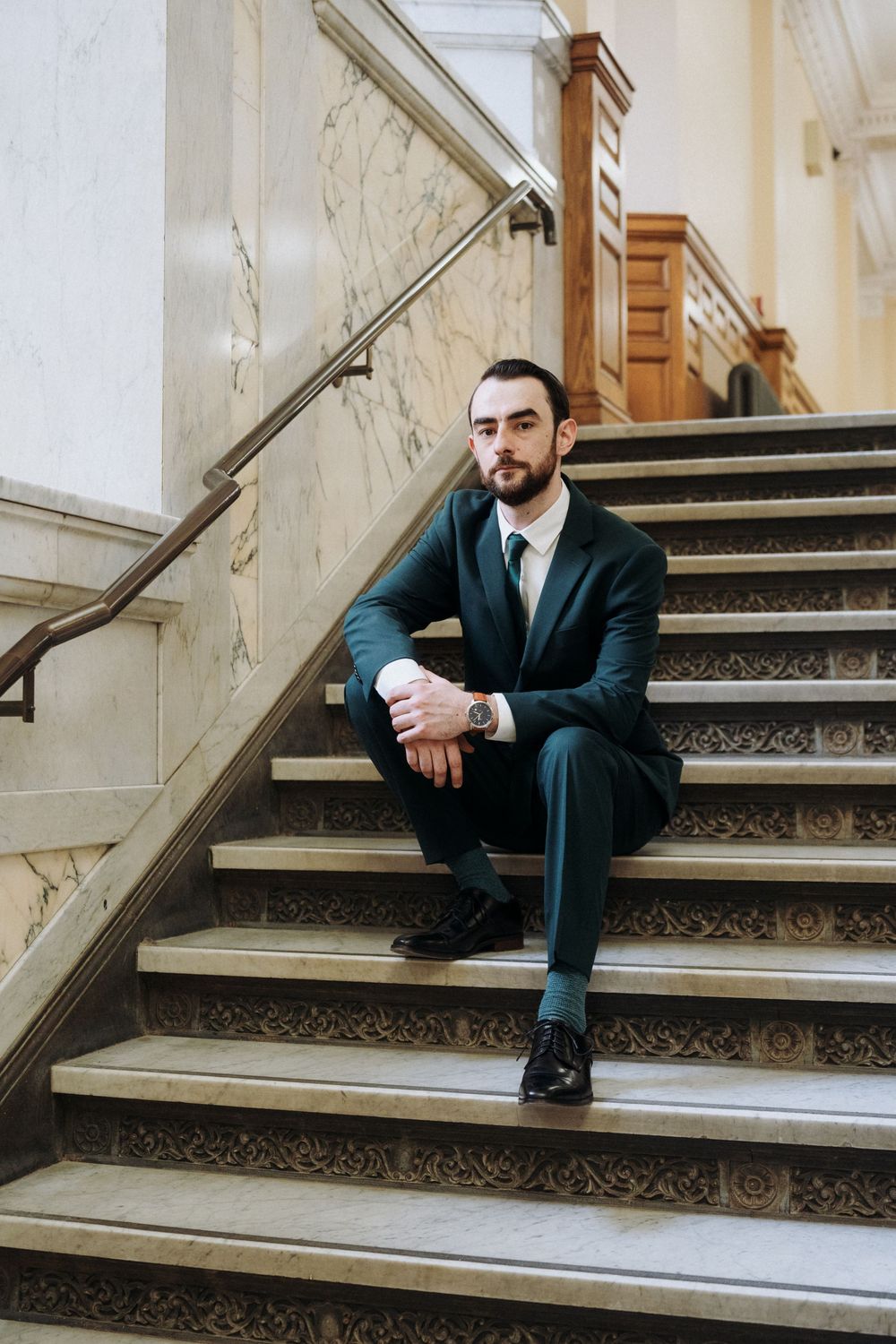 A well-dressed man in a dark green suit sits on ornate marble stairs in an elegant building.
