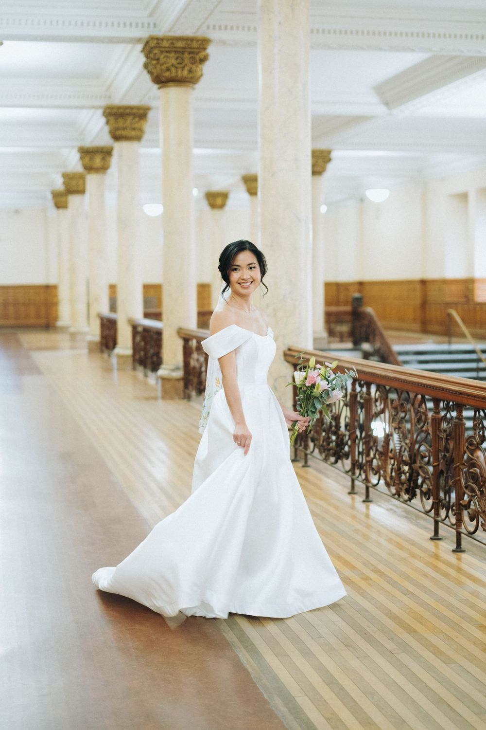 Bride in off-shoulder white gown holds bouquet beside ornate railing in elegant hall with marble columns.