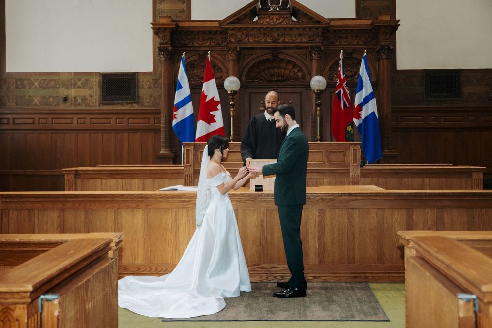 Couple exchanging vows in a wood-paneled courthouse ceremony hall with Canadian flags and an officiant.