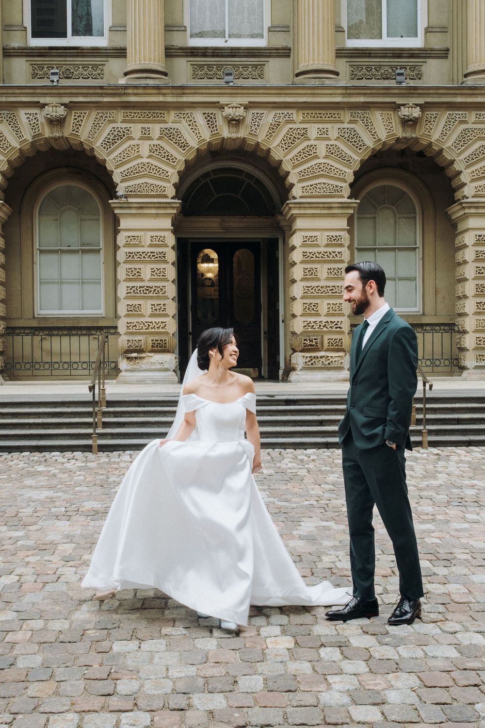 Bride in white off-shoulder gown and groom in dark suit walk on cobblestones before a grand historic building.