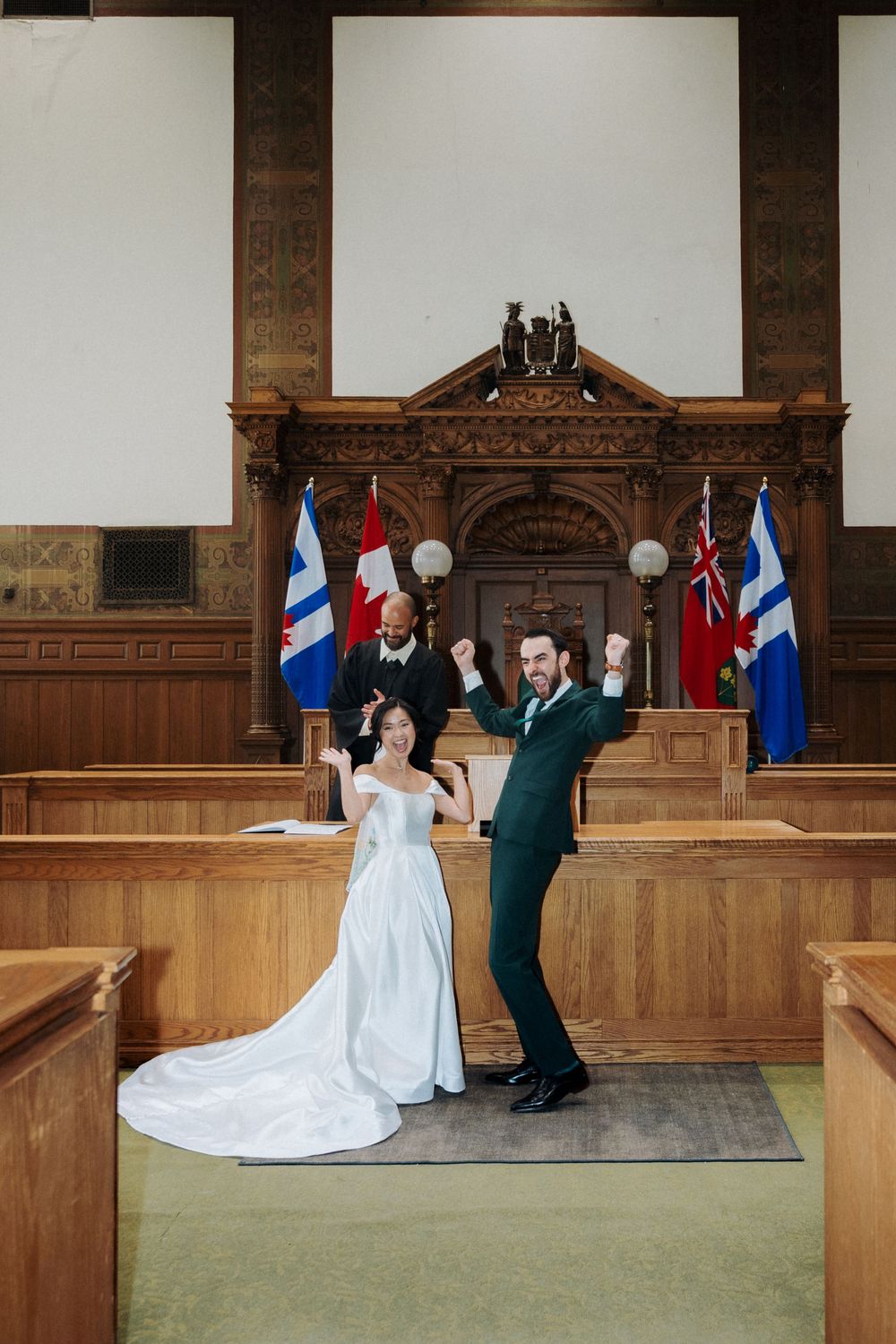 Joyful newlyweds celebrate after courthouse wedding ceremony with officiant and Canadian flags in ornate wood-paneled room.