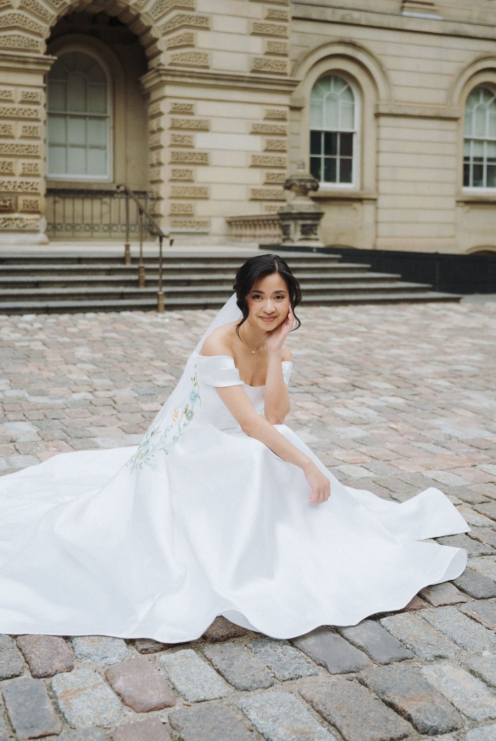 Bride in off-shoulder white gown with floral details sits on cobblestone courtyard before a classical stone building.